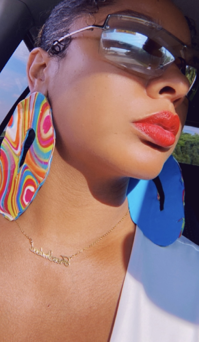 Close-up of a woman wearing sunglasses, colorful earrings, and a necklace, with bright red lipstick and a blue and white outfit.