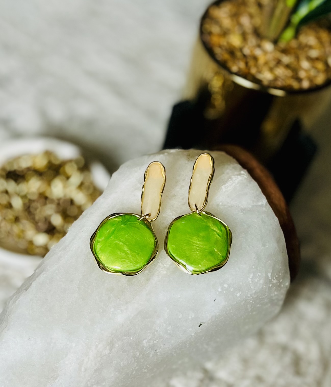 Green gemstone earrings with gold hooks resting on a white surface, with a plant and a bowl in the background.
