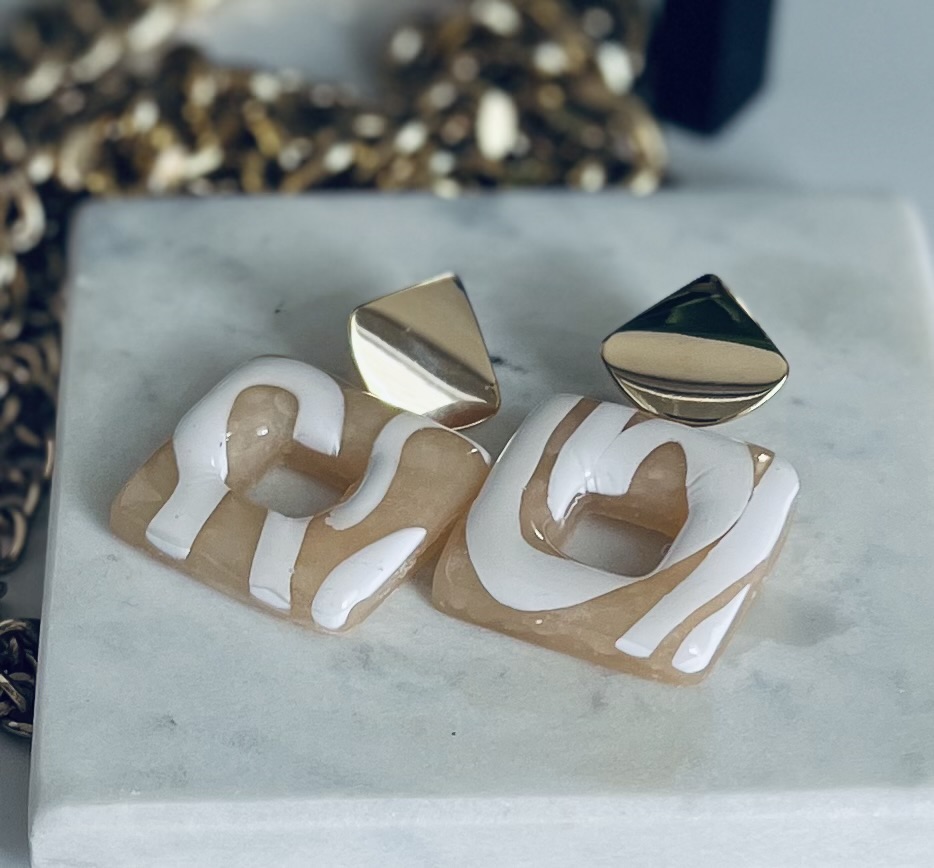 Two gold and white earrings with abstract shapes on a white surface, with a blurred background.