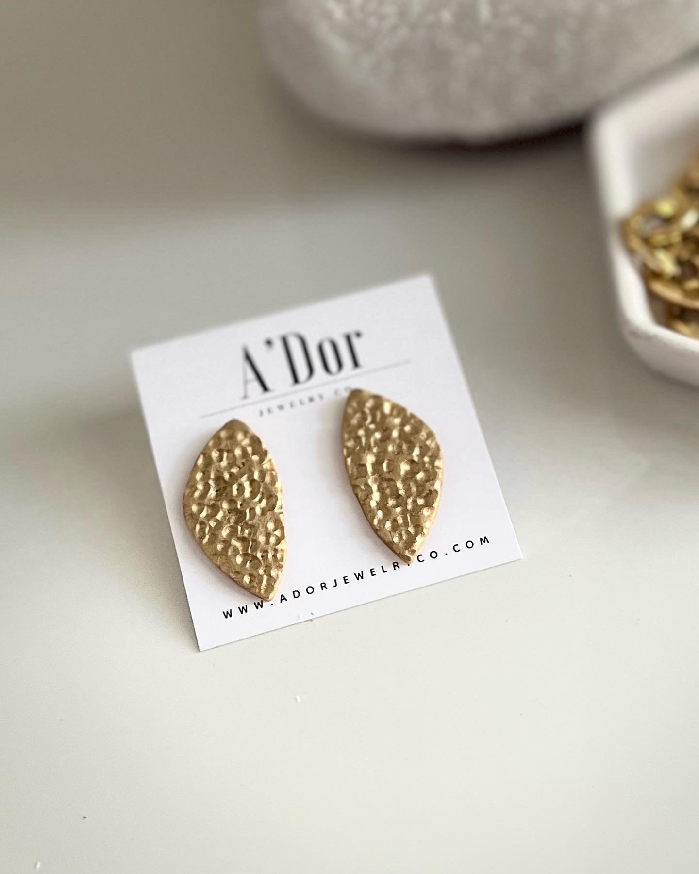 Gold textured oval earrings on a white card with 'A'Dor' branding, placed on a light surface with a bowl of jewelry nearby.