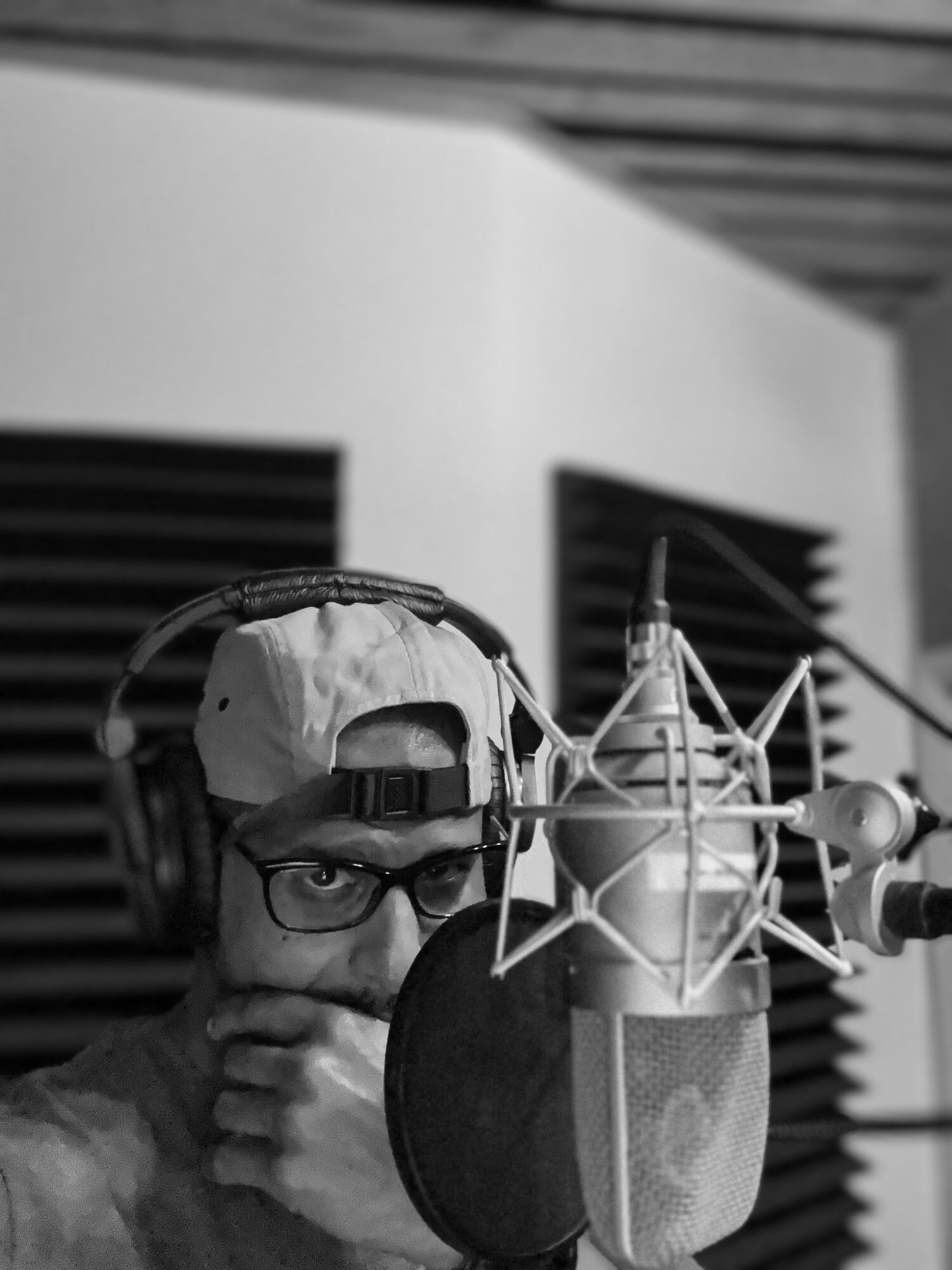Person wearing glasses and a cap, speaking into a large studio microphone with a pop filter, in a recording studio.