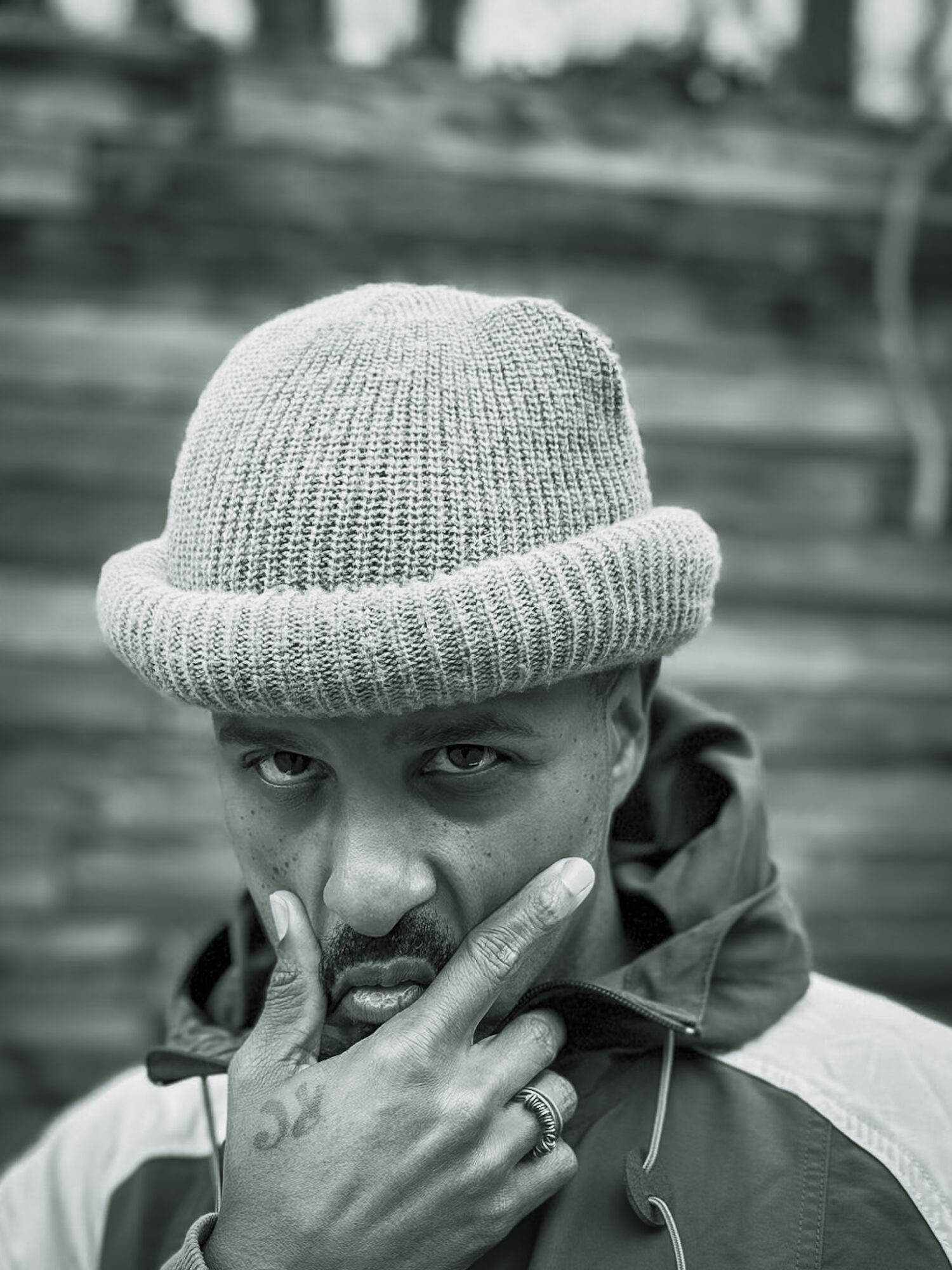 Young man wearing a knit hat, making a peace sign near his face, with a serious expression, outdoors in black and white.