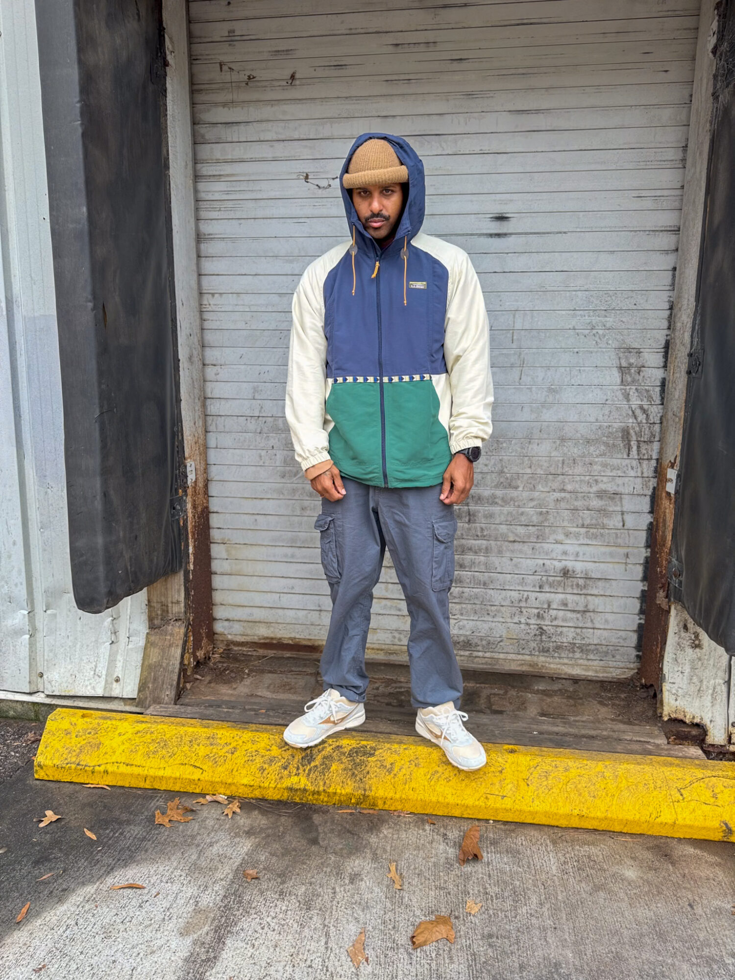 Young man standing outdoors in front of a closed metal shutter, wearing a colorful hooded jacket, gray pants, and white sneakers.