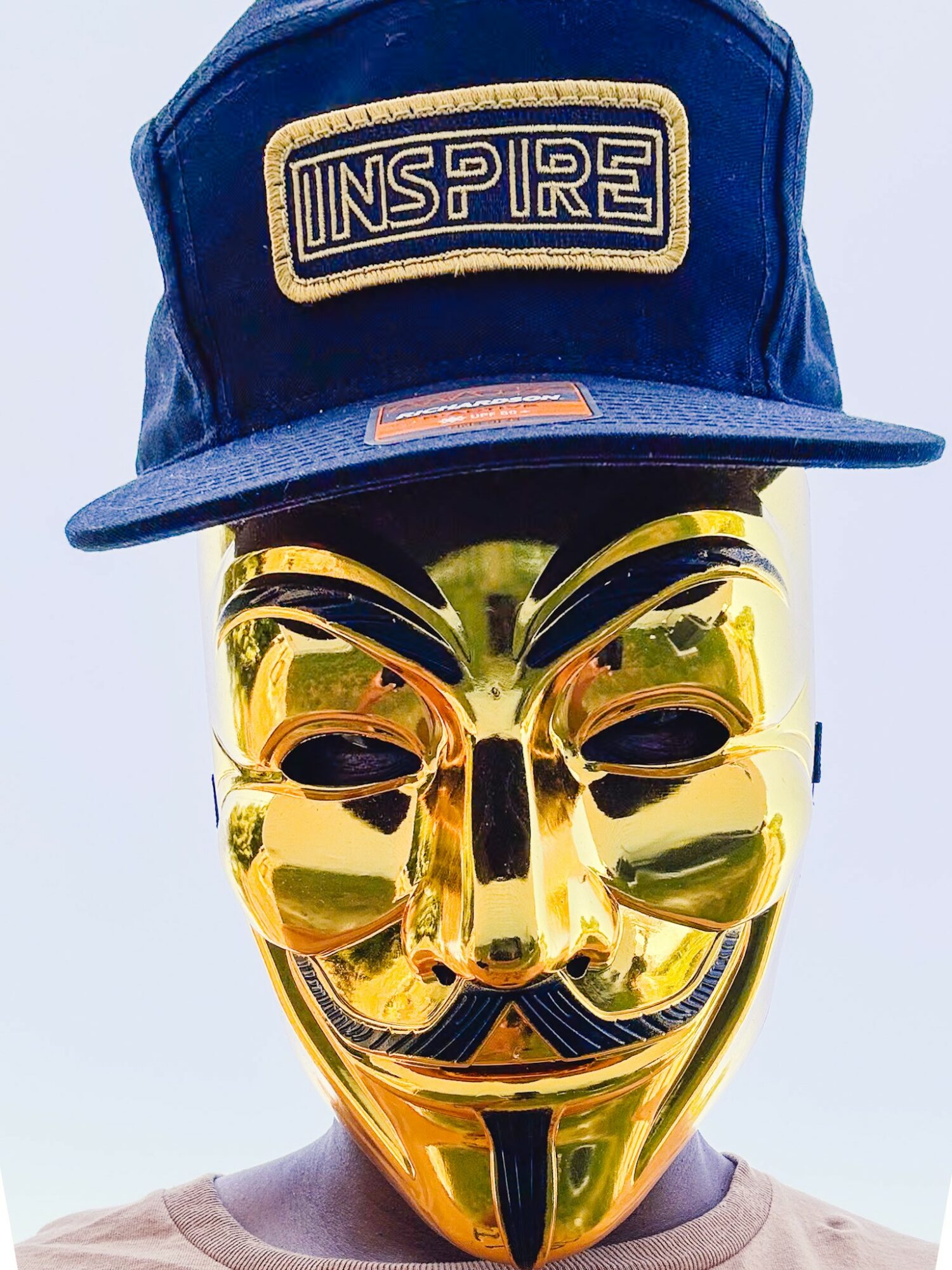 Person wearing a blue cap with 'INSPIRE' patch and a gold Guy Fawkes mask.