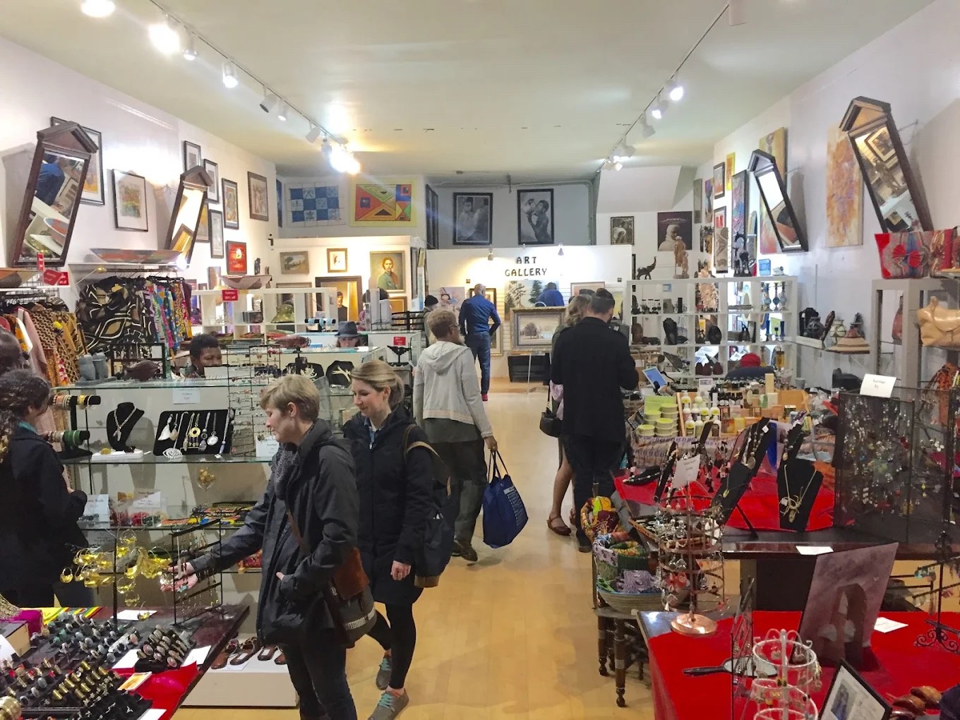 People browsing art and jewelry in an art gallery or store with white walls and various displayed artworks and accessories.