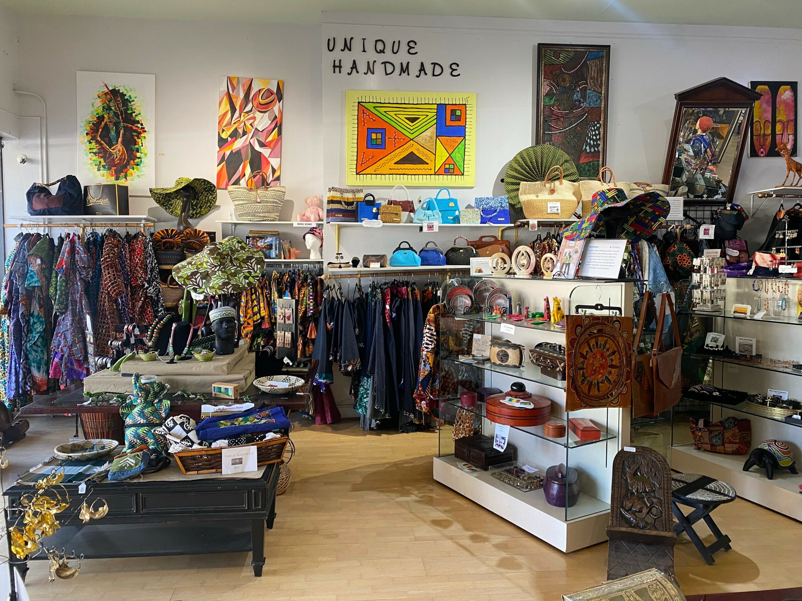 Display of handmade clothing, artwork, and crafts in a store with shelves and racks, colorful decorations, and a sign reading 'Unique Handmade'.