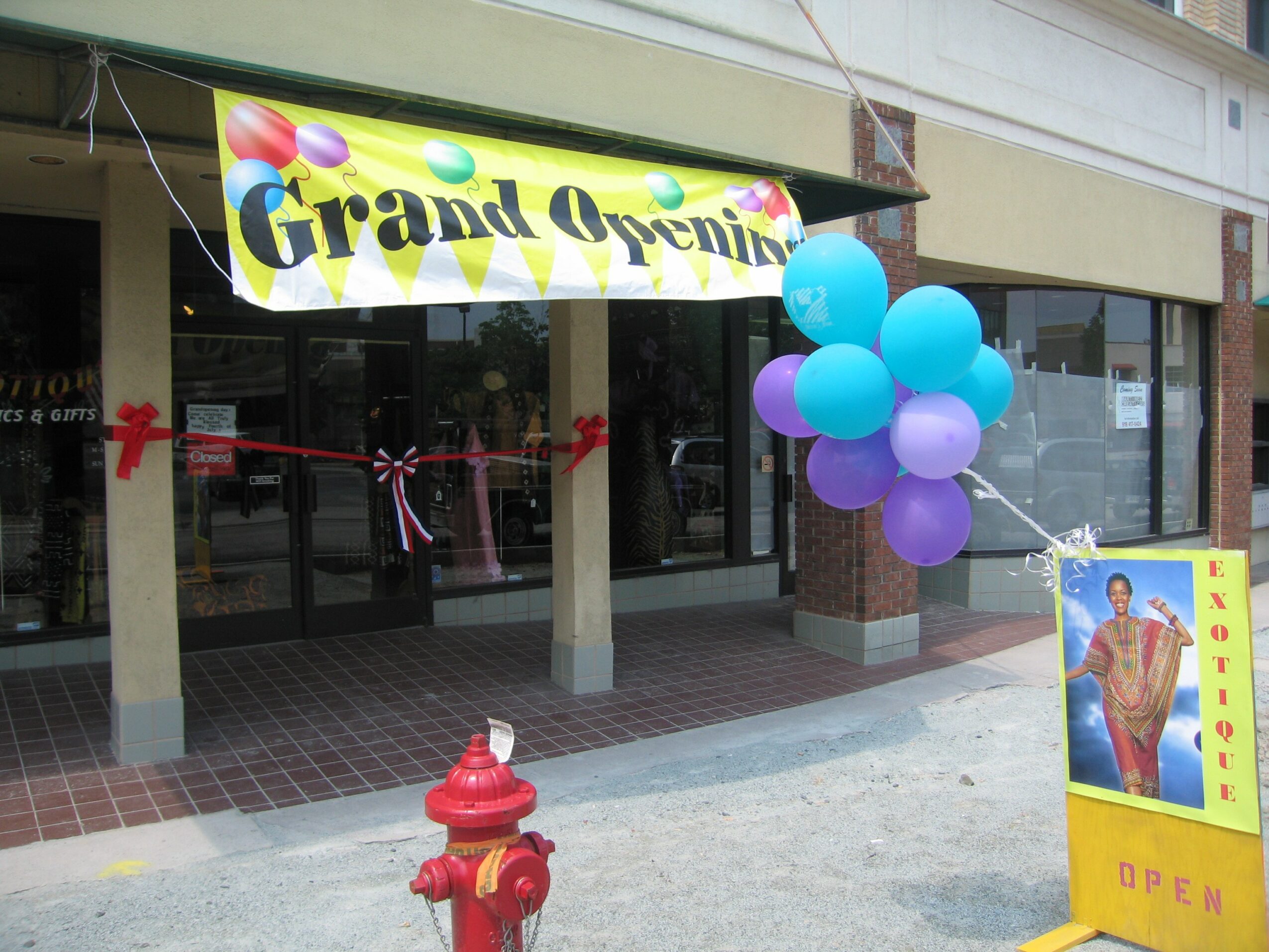 Grand opening banner with balloons outside a storefront, with a promotional poster nearby, and a sidewalk in front.