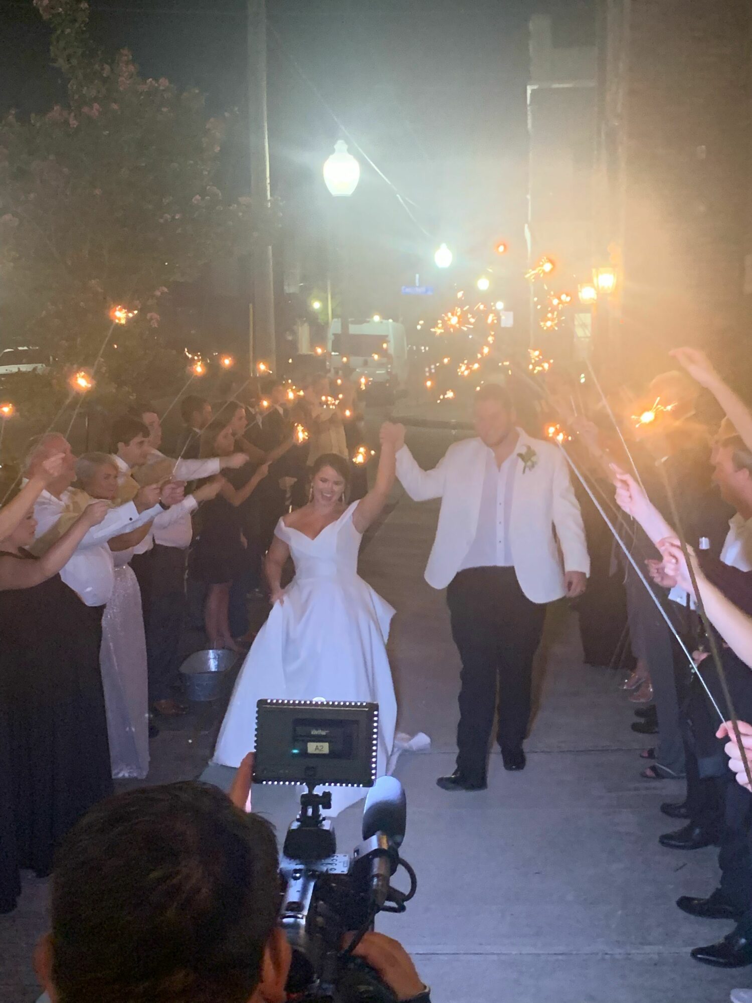 People gather outdoors at night, some holding sparklers, with streetlights and trees in the background.