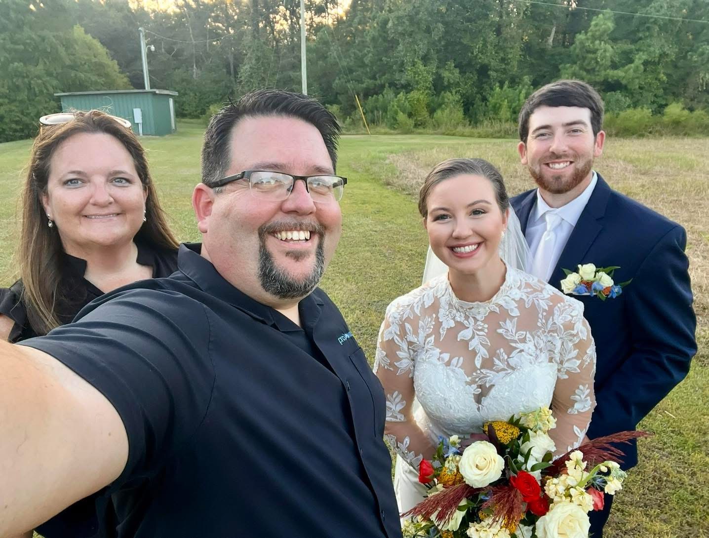 Four people smiling outdoors, one holding a bouquet, with trees and a field in the background.