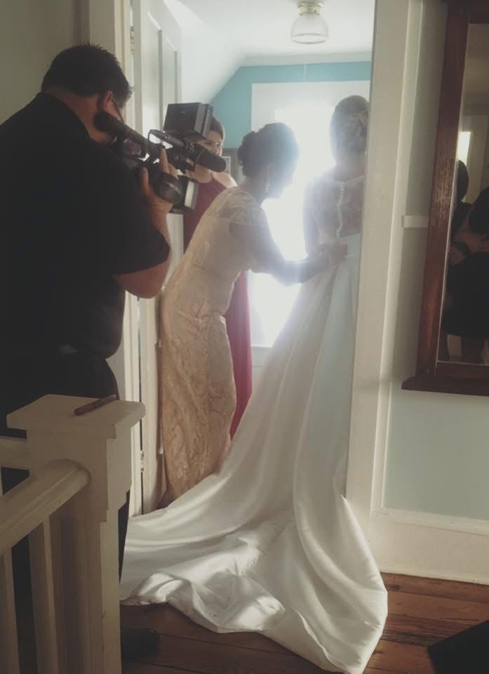 Two women, one in a wedding dress, hold hands in a brightly lit room, with a photographer capturing the moment.