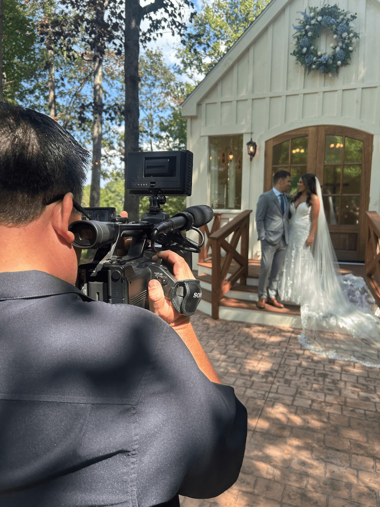Person filming a wedding outside a white building with a wreath, couple standing near the entrance, surrounded by trees.