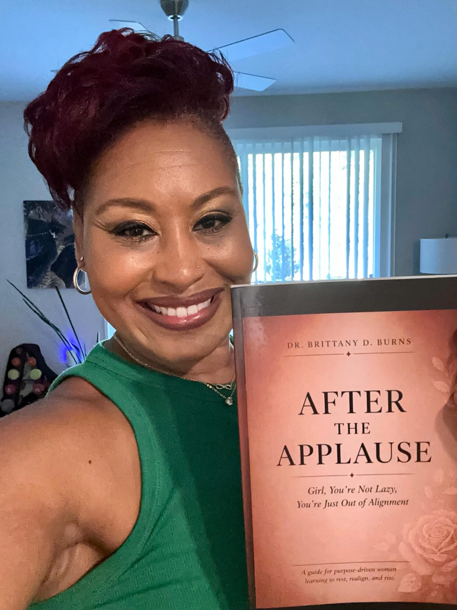Smiling woman with short red hair in green top holding a book titled 'After the Applause' in a room with window blinds.