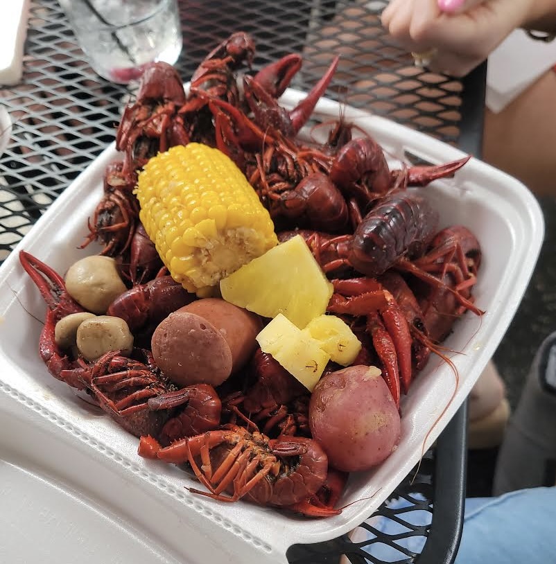 Plate with cooked crawfish, corn on the cob, potatoes, and lemon wedge on a table, with a glass and a hand nearby.