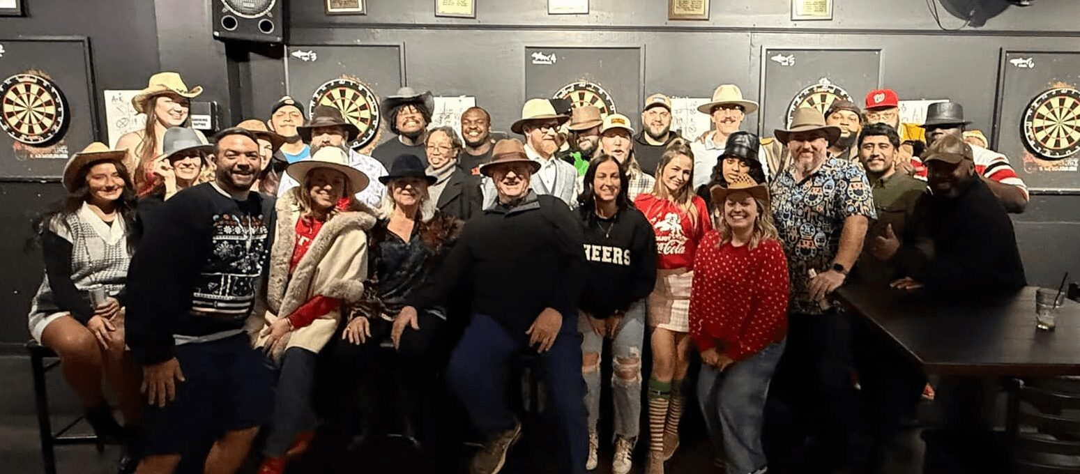 Group of people wearing cowboy hats in a room with dartboards on the wall, smiling and posing for a photo.