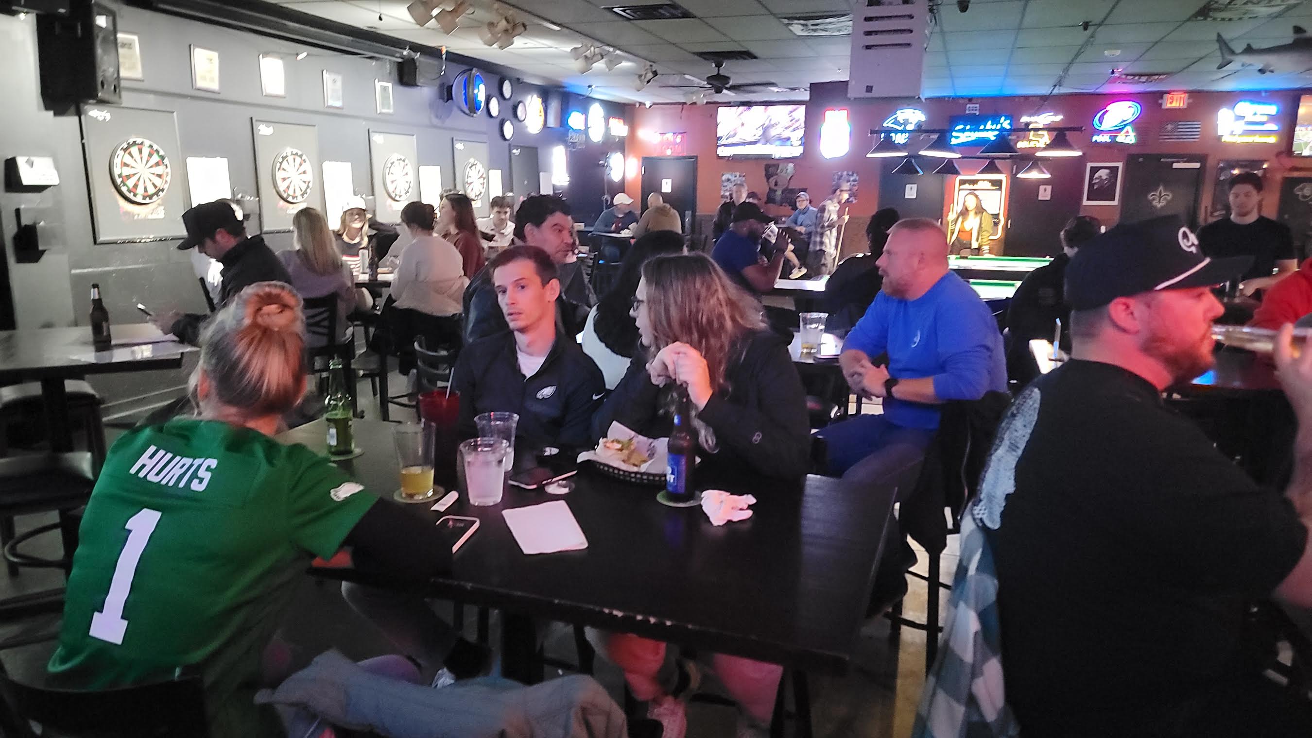 People sitting at tables in a bar or pub, some drinking and socializing, with dartboards on the wall, dim lighting.