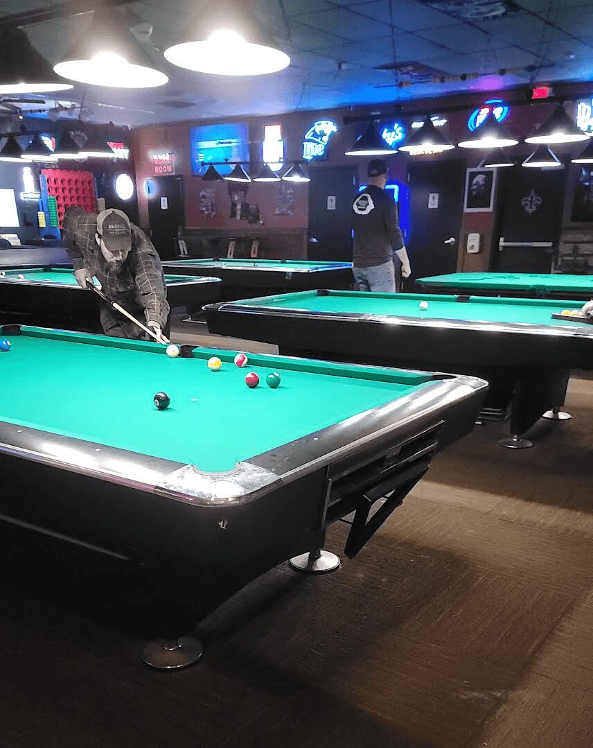 Pool table with balls, people playing, neon signs, and overhead lights in a game room.