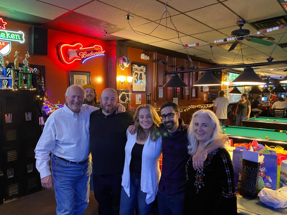 Group of five people smiling in a bar with neon signs and pool tables, some with arms around each other.