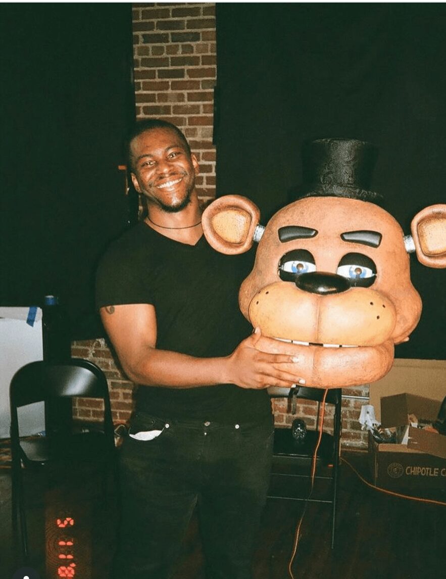 Person smiling holding a large bear head costume mask, brick wall background, indoor setting