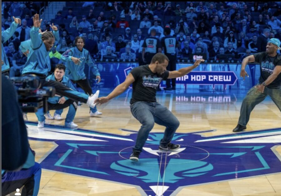 People dancing on a basketball court with a crowd in the background, some wearing teal shirts, and a sign reading 'HIP HOP CREW'.