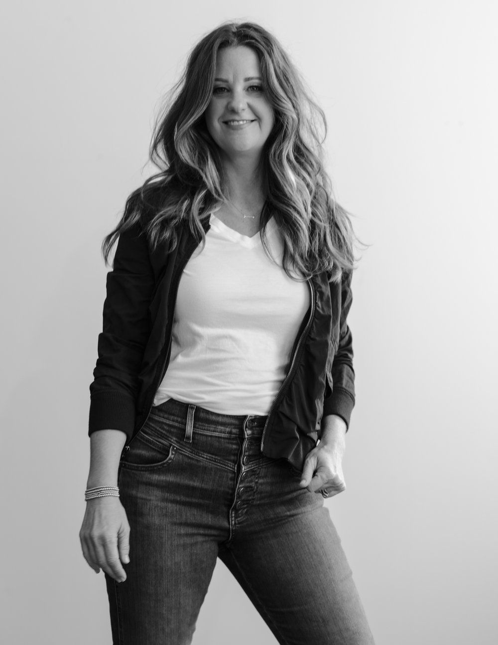 Woman with long wavy hair smiling, wearing a white shirt and dark jeans, standing against a plain background.