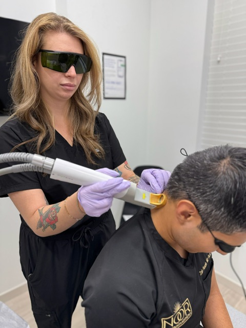 A woman wearing protective glasses and gloves performs a laser treatment on a man's face in a clinical setting.