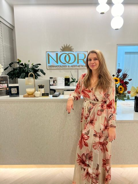 Woman in floral dress standing in front of a reception desk with flowers and decor, smiling, indoors.