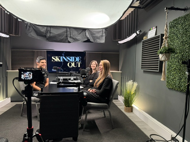 Three women sit at a table in a recording studio with a camera recording them, and a sign reads 'SKIN SIDE OUT'.