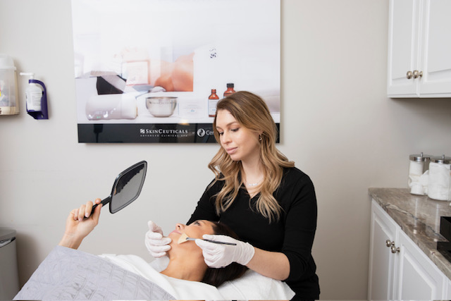 A woman in a black top performs a facial treatment on a client in a clinic room.