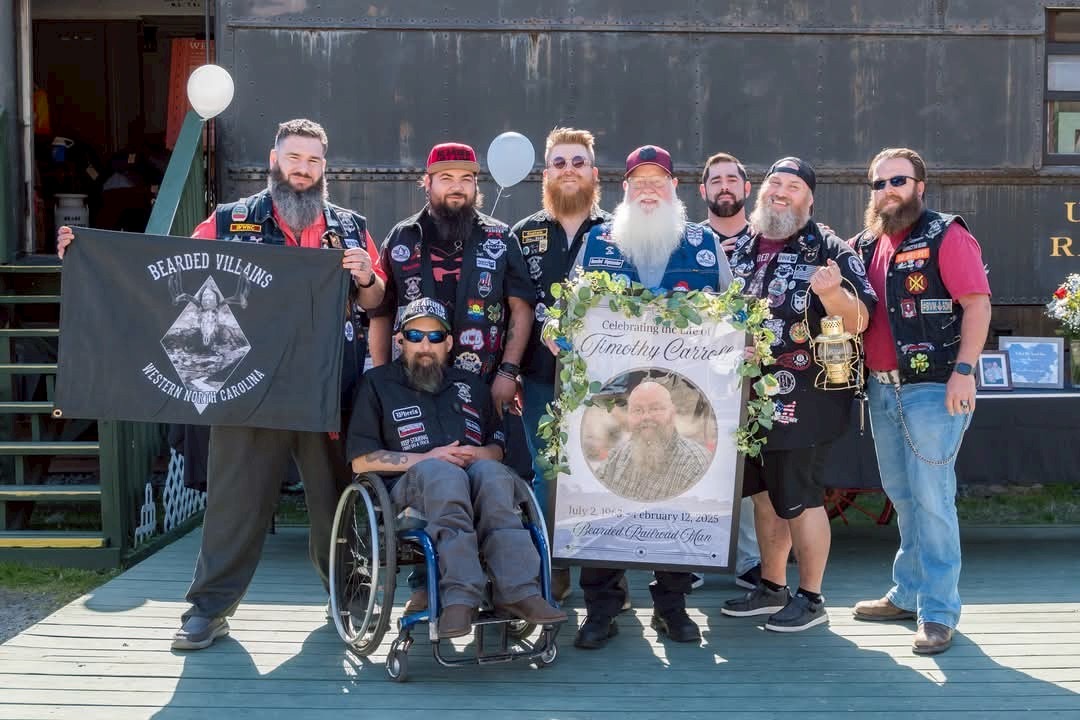 Group of eight men, one in wheelchair, holding a floral wreath, standing outdoors in front of a building, some wearing vests with patches.