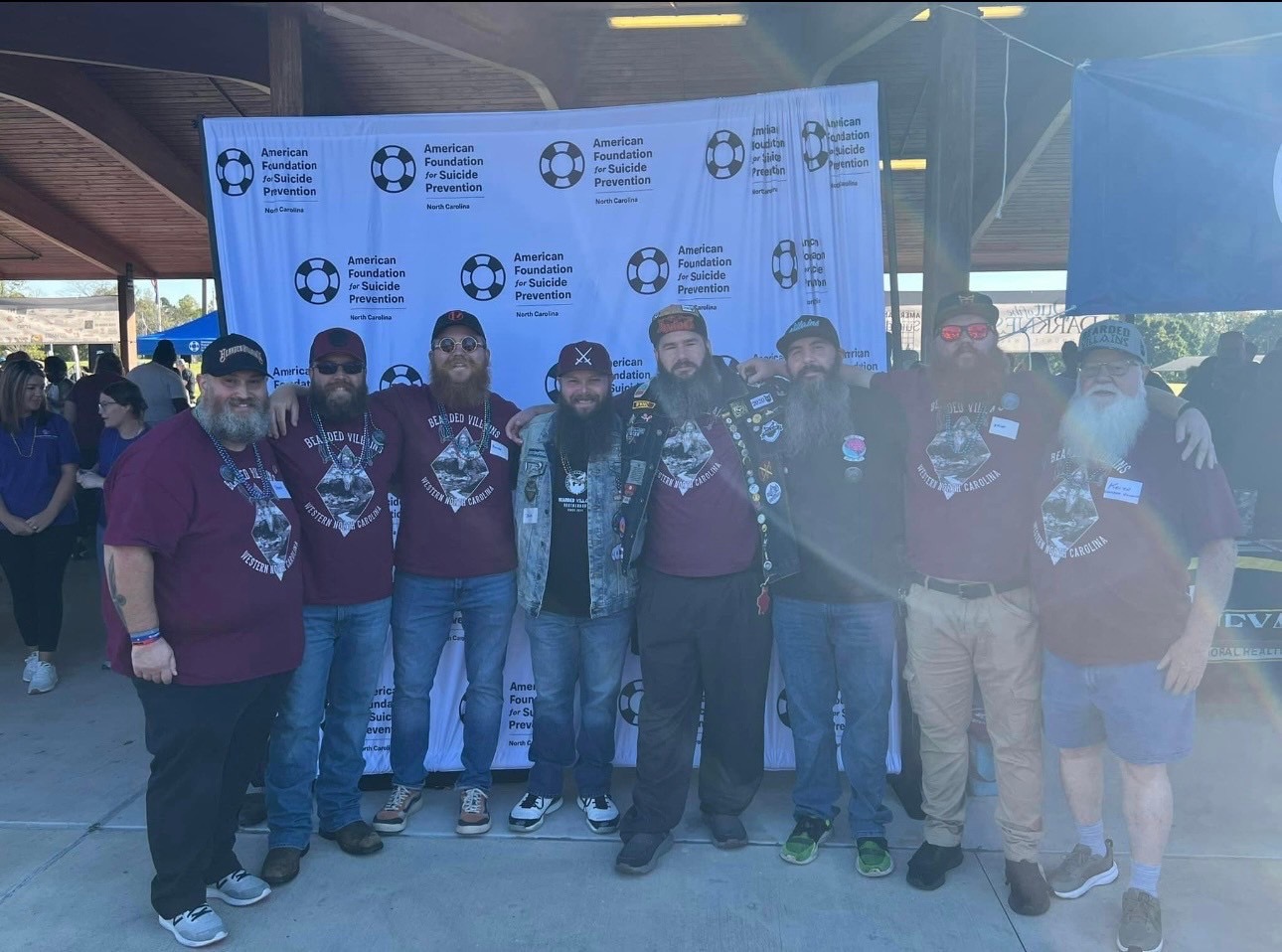Group of people standing in front of a blue backdrop with logos, outdoors, some wearing maroon shirts, some with masks.