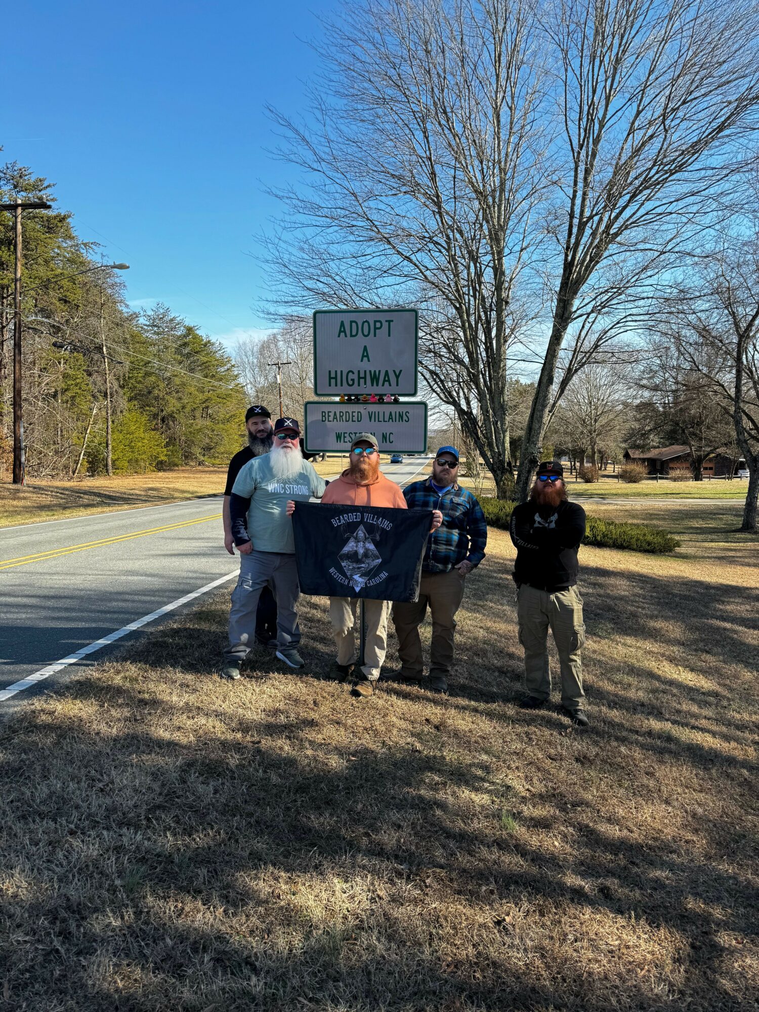 Group of people standing near a road sign, trees, and a clear blue sky, with shadows on the ground.