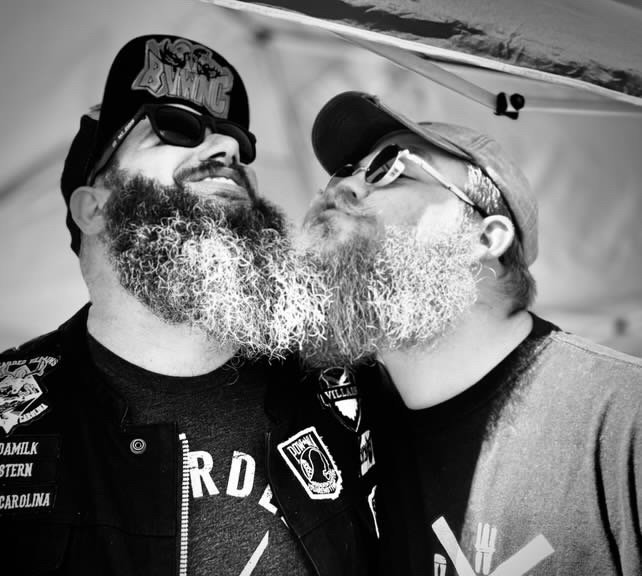 Two men with beards and sunglasses face each other, smiling and close together outdoors, with a tent in the background.