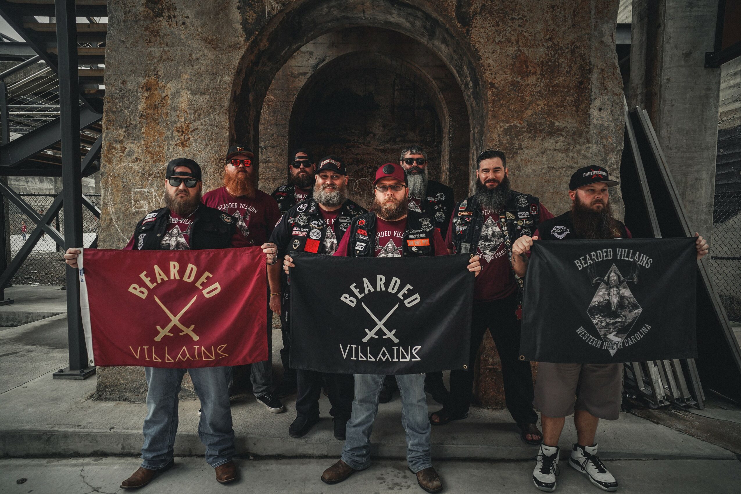 Group of eight men standing in front of an arched brick wall, holding three flags with logos and text.