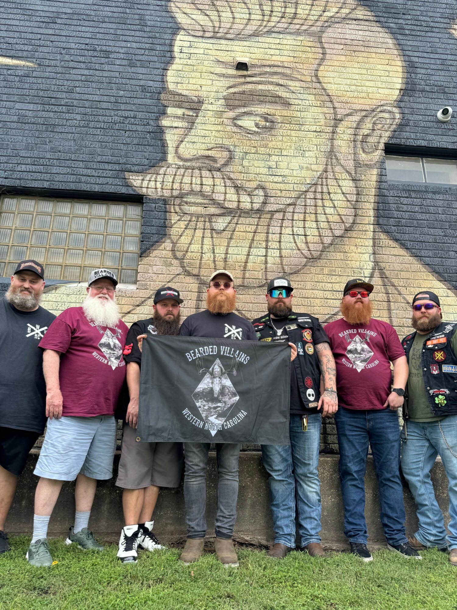 Group of nine people standing outdoors in front of a large mural of a face on a brick wall, some holding a black flag.
