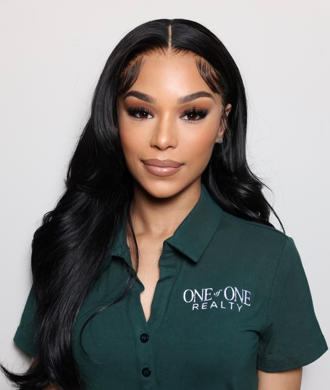 Woman with long black hair wearing a green shirt with 'ONE & ONE REALTY' logo, looking at camera.