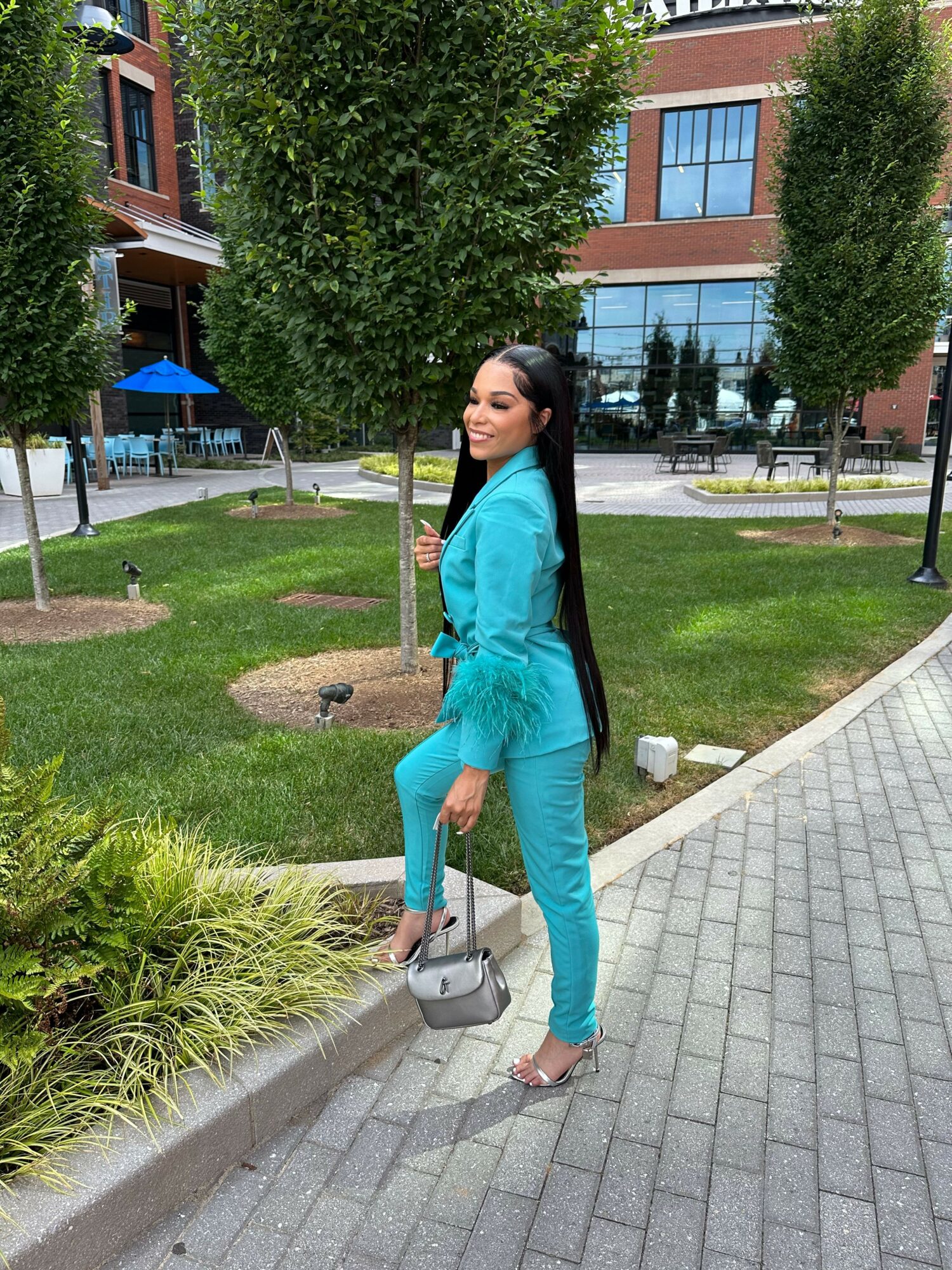 Woman in turquoise outfit walking on sidewalk with greenery and buildings in background.