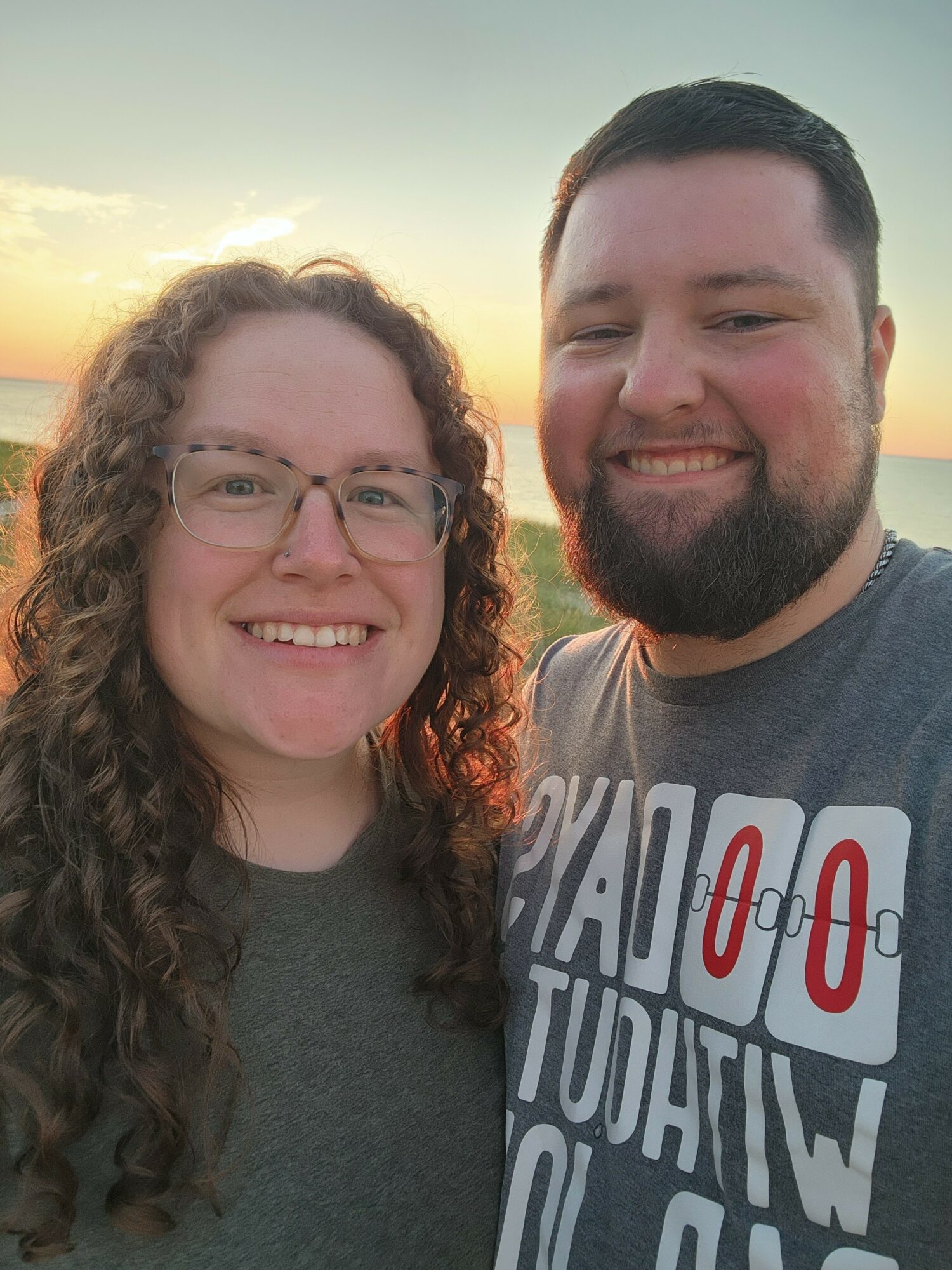 Two people smiling outdoors at sunset, one with curly hair and glasses, the other with short dark hair and a beard.
