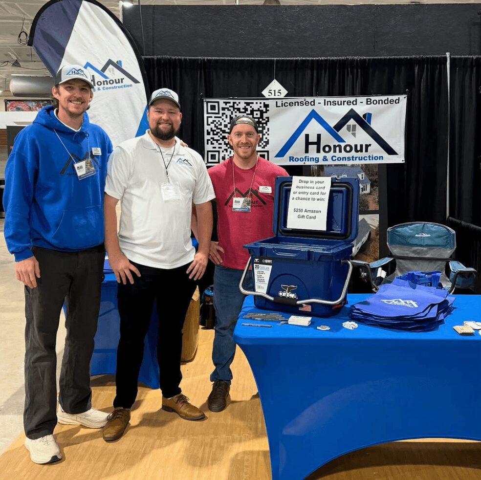 Three men stand in front of a booth with a sign reading 'Honour Roofing & Construction,' smiling. The booth has a blue tablecloth and various items displayed.