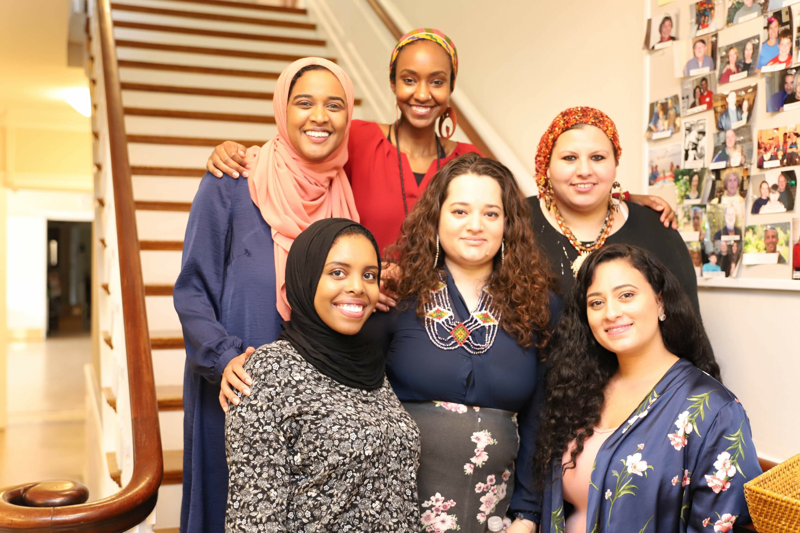 Six women standing together indoors, smiling, with a staircase and photo collage in the background.