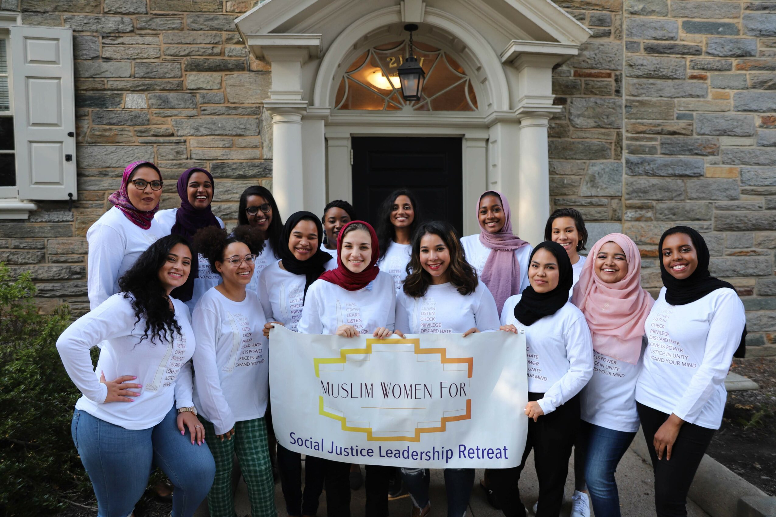 Group of women standing outside a stone building holding a banner for a social justice retreat, smiling and wearing white tops.