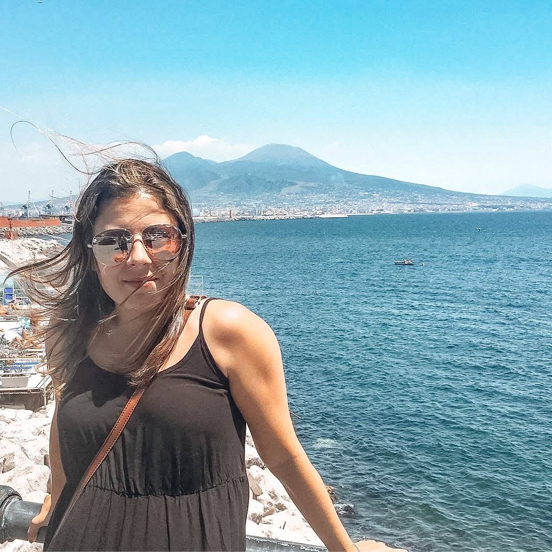 Woman with sunglasses and a black dress stands by the sea with a mountain in the background.