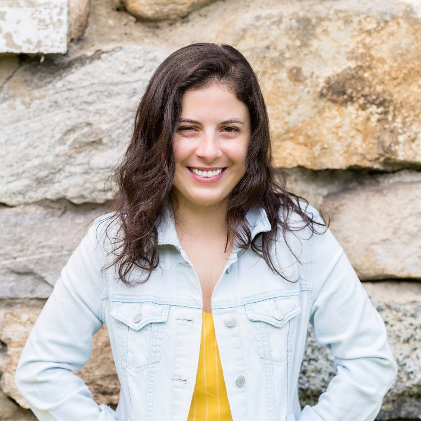 Young woman with dark wavy hair smiling, wearing a light denim jacket and yellow top, standing outdoors against a stone wall.