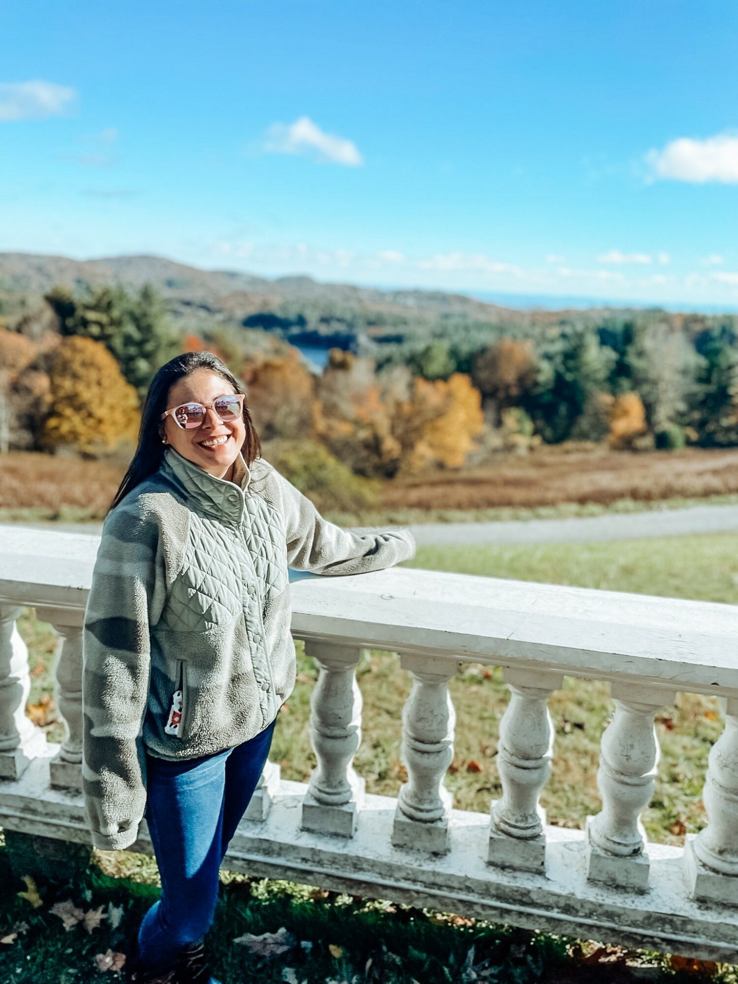 Smiling woman with glasses leaning on a white railing outdoors, with trees and a blue sky in the background.