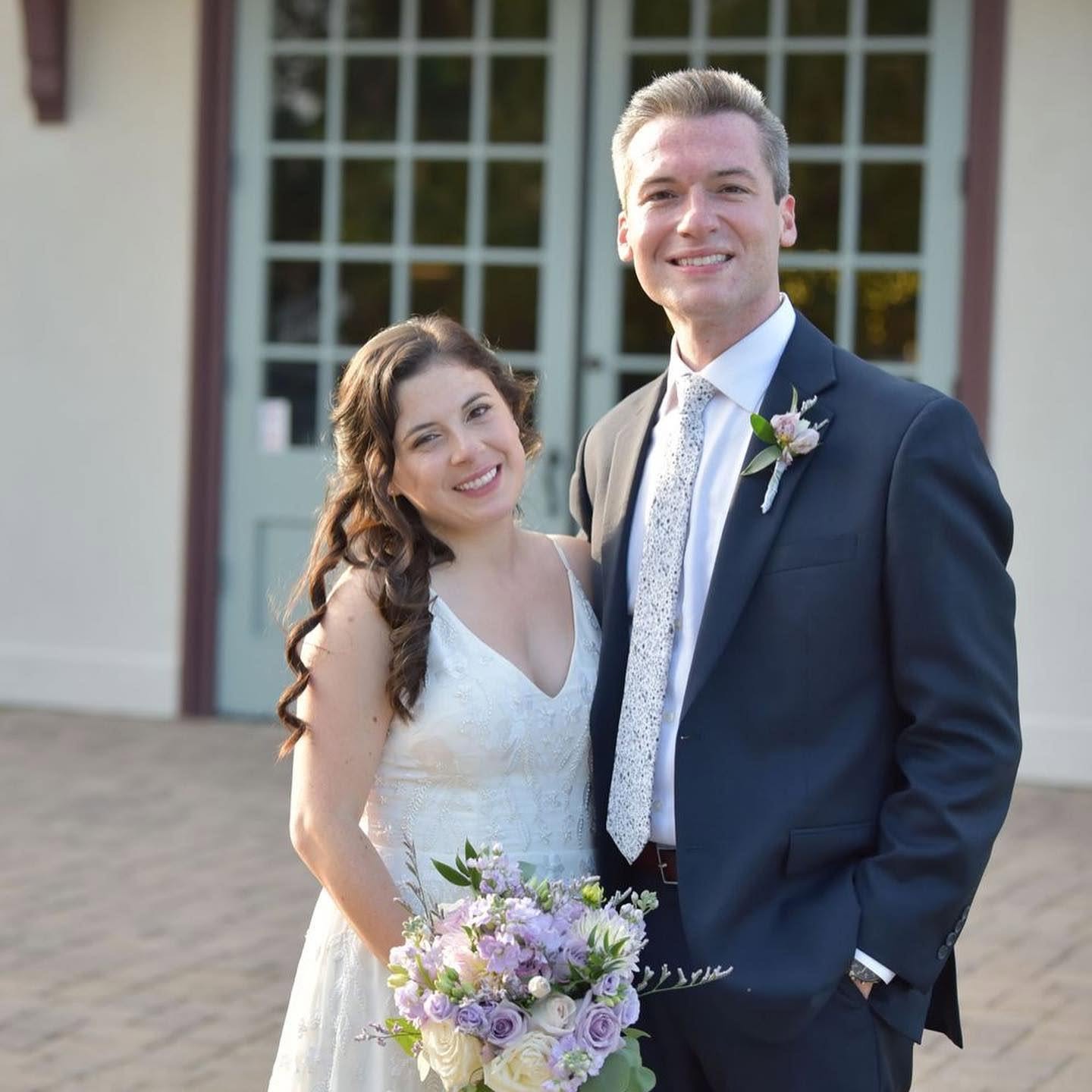A smiling bride and groom stand outdoors in front of a building with large windows, holding a bouquet of flowers.