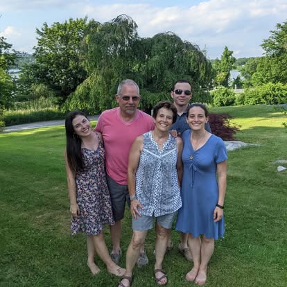 Group of five people standing outdoors on grass with trees and sky in background, smiling at the camera.