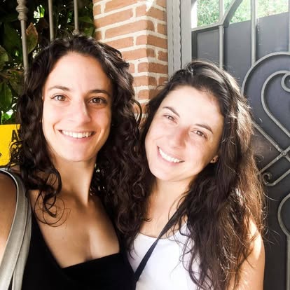 Two women with long, dark, curly hair smiling outdoors near a brick wall and metal gate.