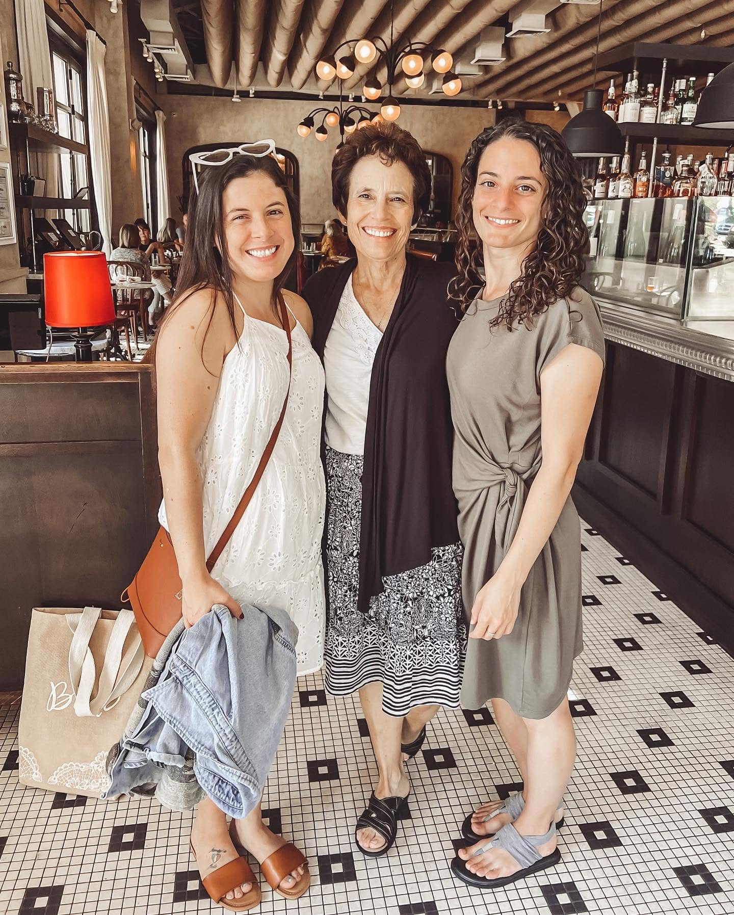 Three women standing together inside a cafe, smiling, with a bar and shelves in the background.