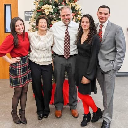Group of five people standing in front of a decorated Christmas tree, smiling, dressed in festive and formal clothing.