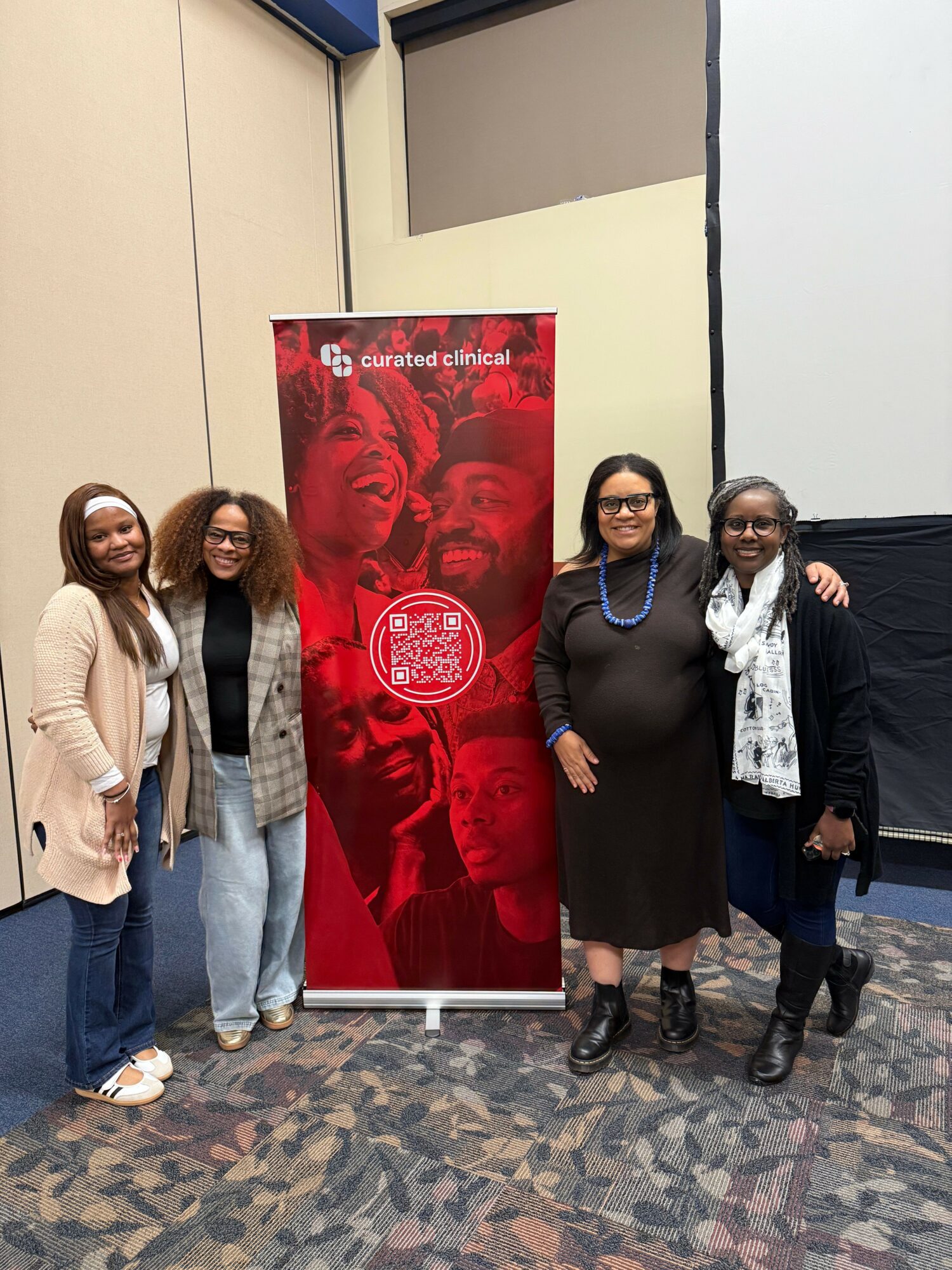 Four women standing in front of a red and black banner with a QR code, in a room with patterned carpet.