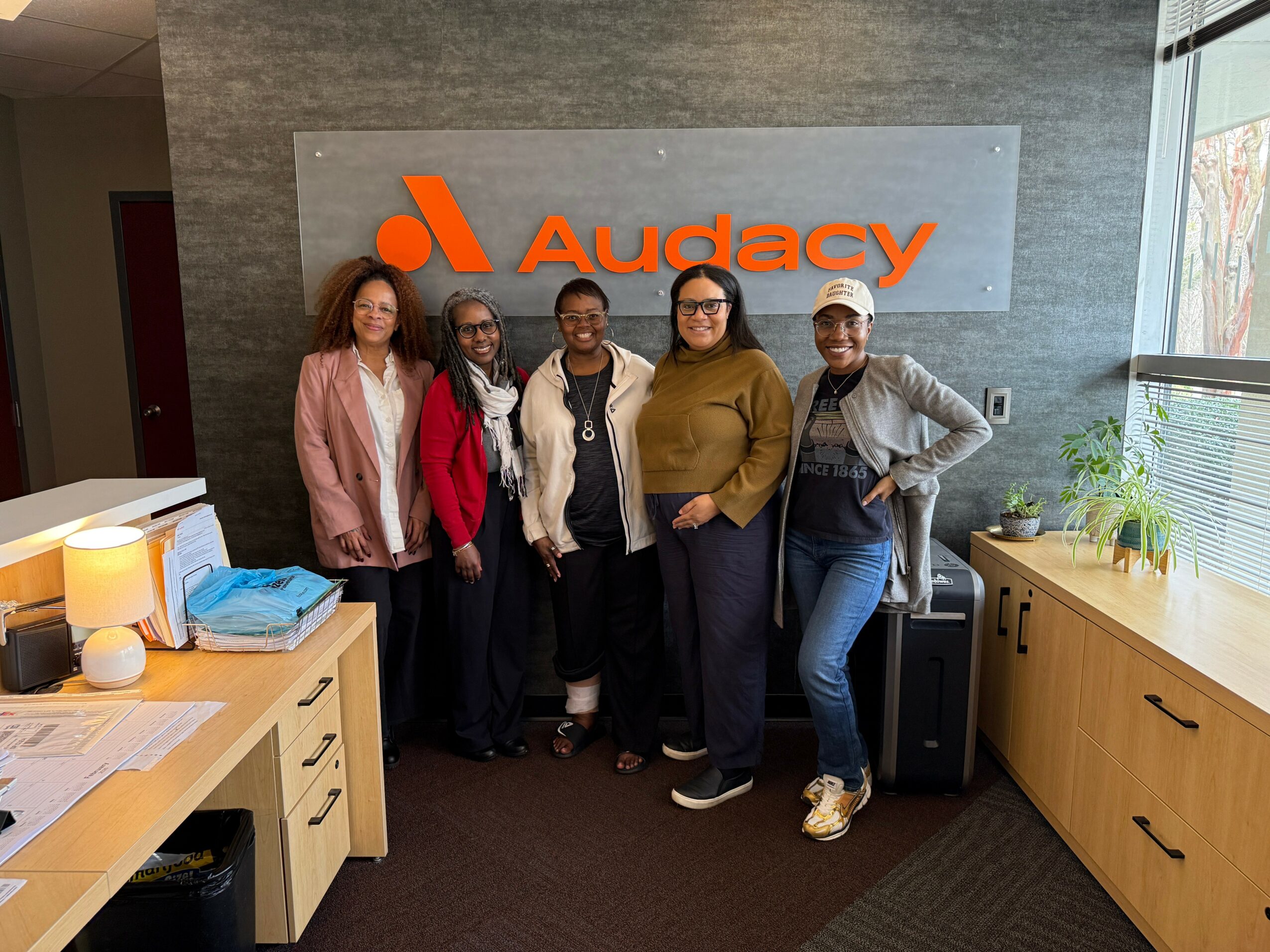 Five women stand together in front of a wall with an Audacy sign, smiling indoors with a window and plants nearby.